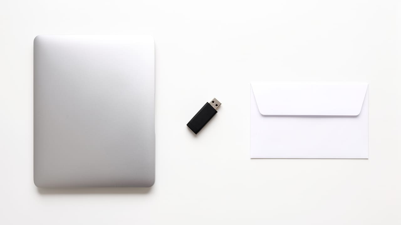 Flat-lay of a closed laptop, a small USB drive, and a plain sealed envelope arranged on a clean white surface